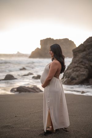 Pregnant woman in white dress stands on beach at sunset with rocky coastline in background.