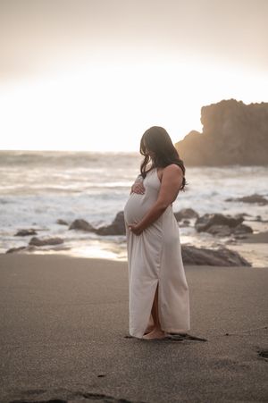 A woman in a flowing white dress stands on a beach at sunset, cradling her pregnant belly.