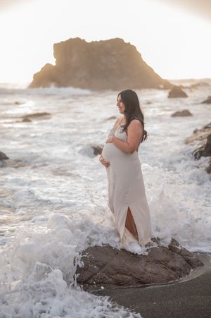 Pregnant woman in flowing white dress stands on rocky beach during sunset as waves crash around her at dramatic coastline.