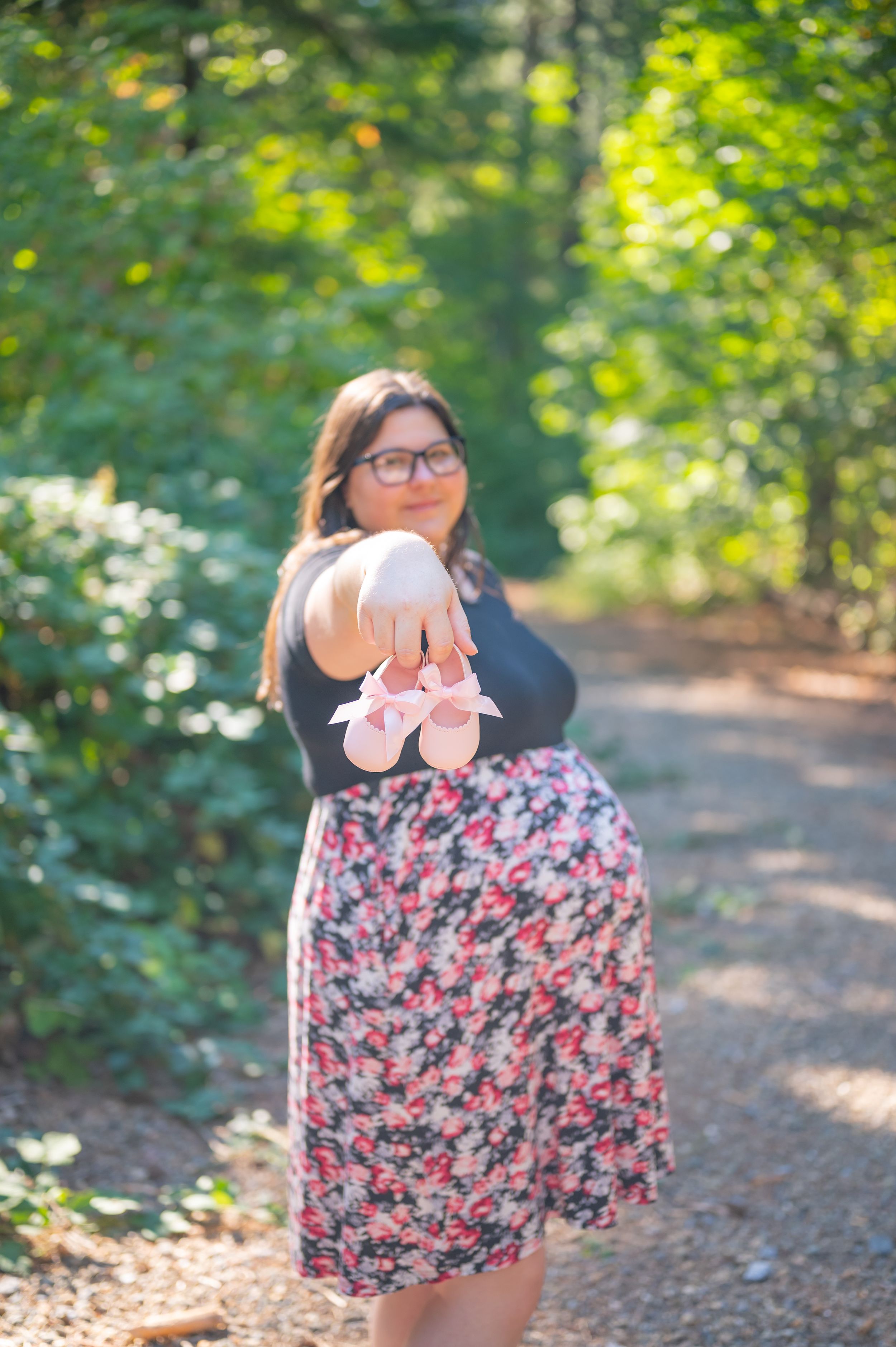 A person in a floral dress holding tiny pink baby shoes in a sunlit wooded path.