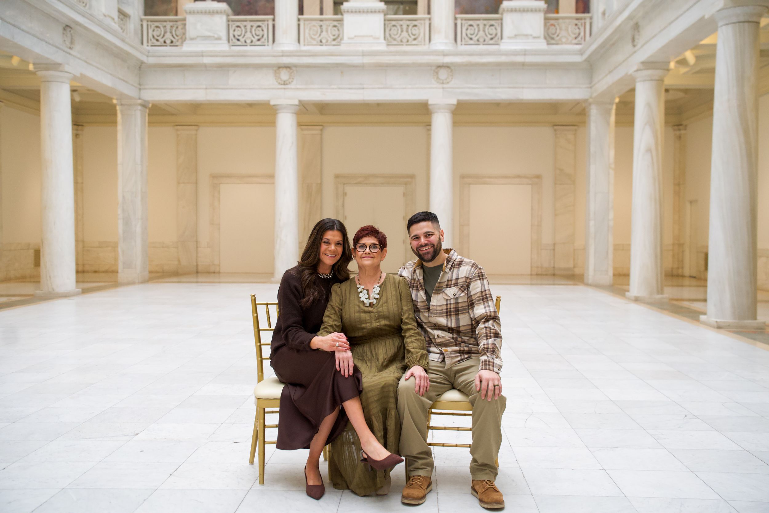 Three people posing together in an elegant columned hall with marble floors and classical architecture.