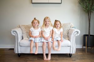 Three young blonde girls in matching white tops and skirts sitting together on a beige sofa, smiling.