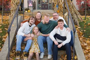 Family group sits together on outdoor stone steps surrounded by fallen autumn leaves in park setting.