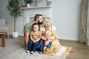 A family sits together on hardwood floor in a bright minimalist living room with white walls and indoor plants.