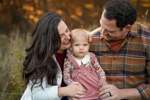 A family shares a tender moment outdoors during autumn with their baby wearing a pink jumper dress.