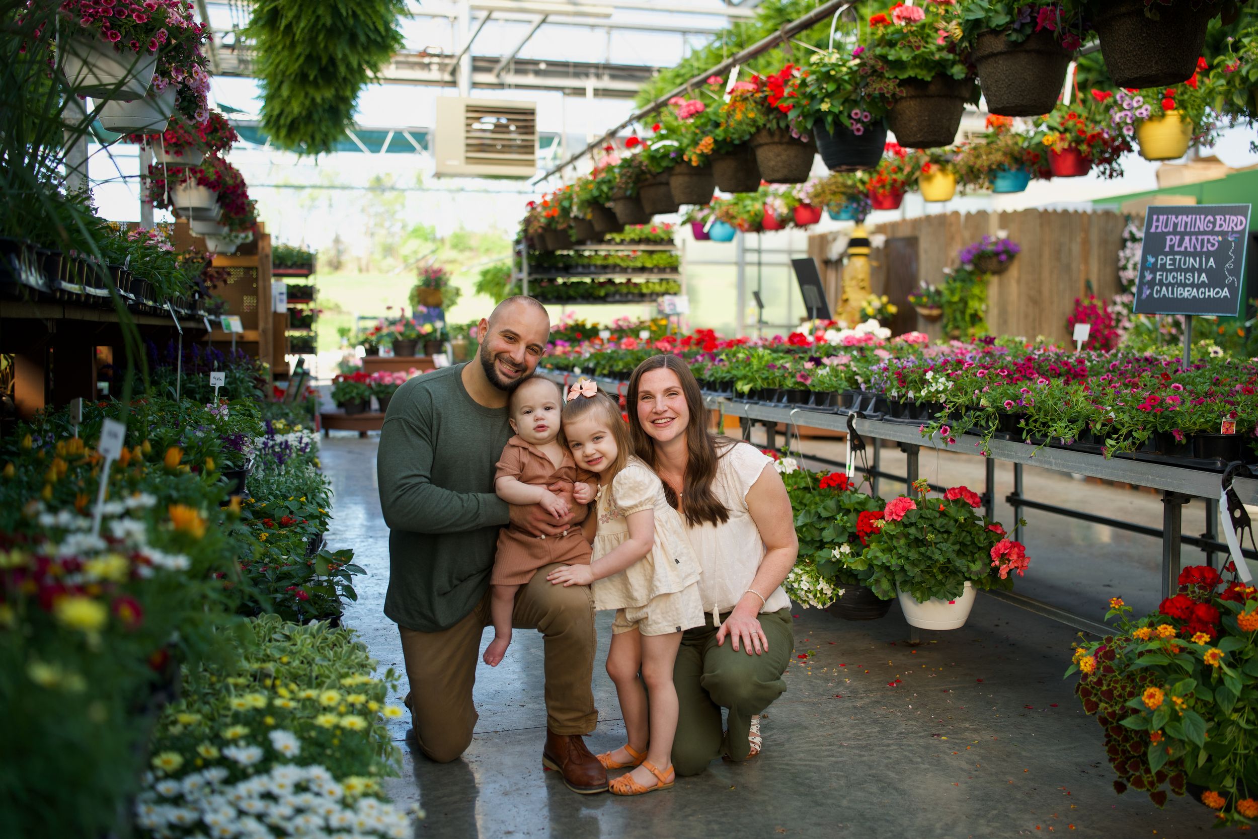 Happy family of four posing together in a colorful garden center surrounded by vibrant flowering plants.
