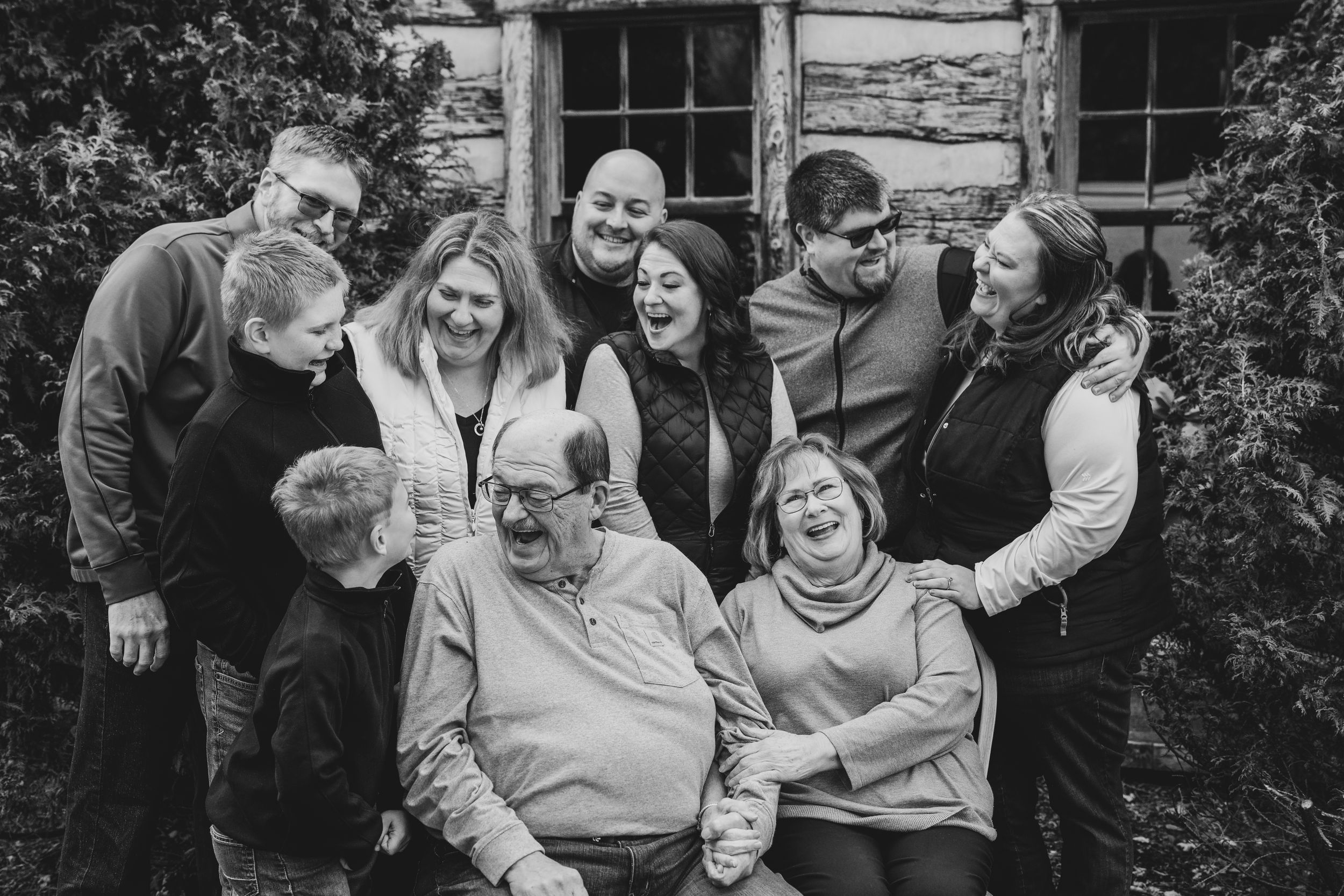 Black and white family portrait of multigenerational group laughing together outside a rustic log cabin.