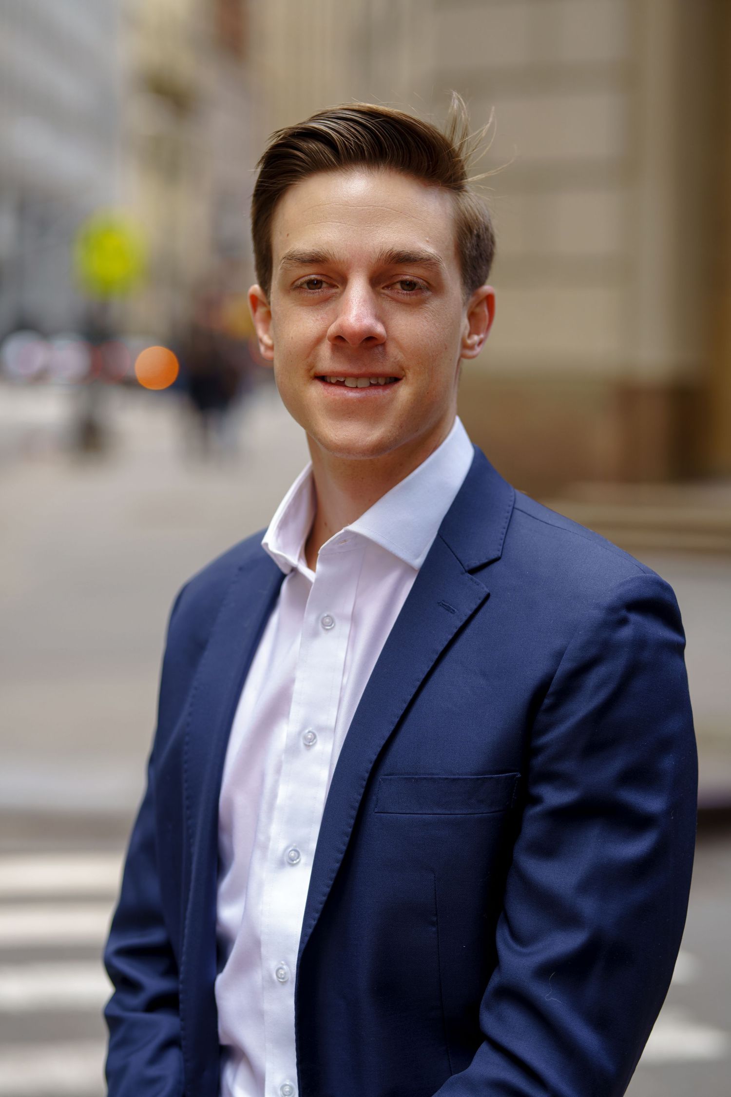A professional headshot of a young executive in a navy suit on an urban street.