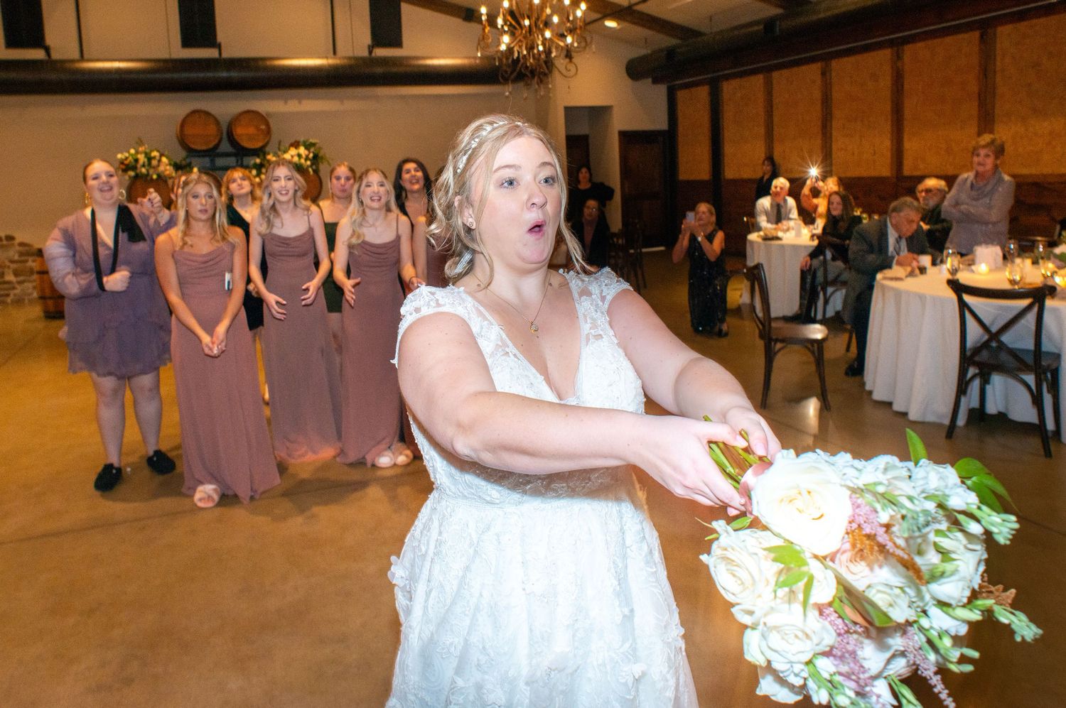 Group in purple bridesmaid dresses watches as someone tosses a wedding bouquet during a rustic indoor reception.