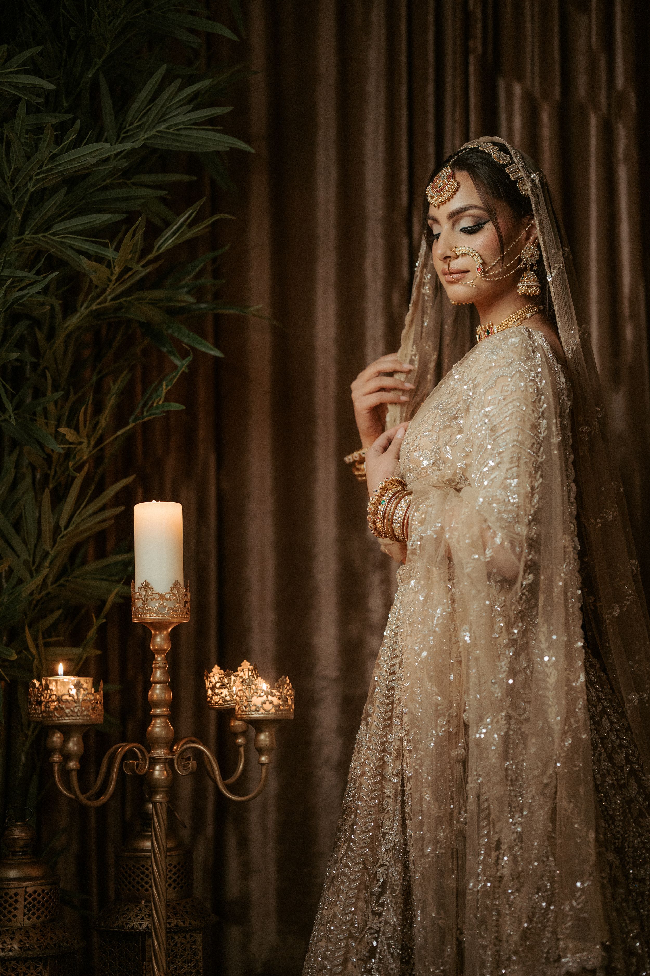 Elegant bride in embellished beige lehenga poses near lit candles in romantic setting.