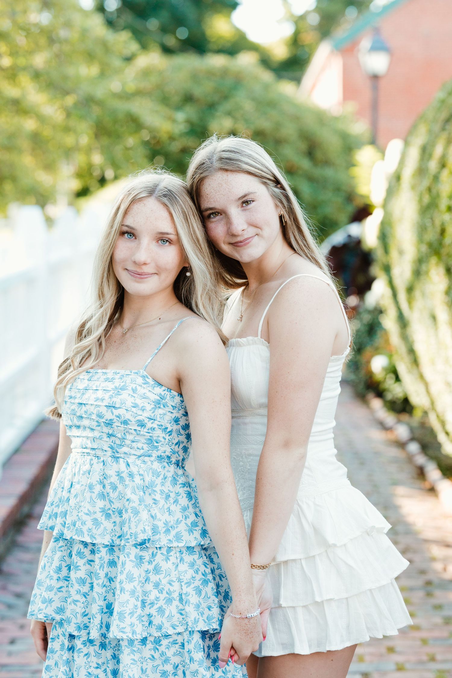 Two young women in summer dresses, one wearing blue floral and one in white ruffled, pose together on a walkway.