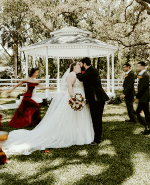 A wedding ceremony takes place under a white gazebo surrounded by trees.