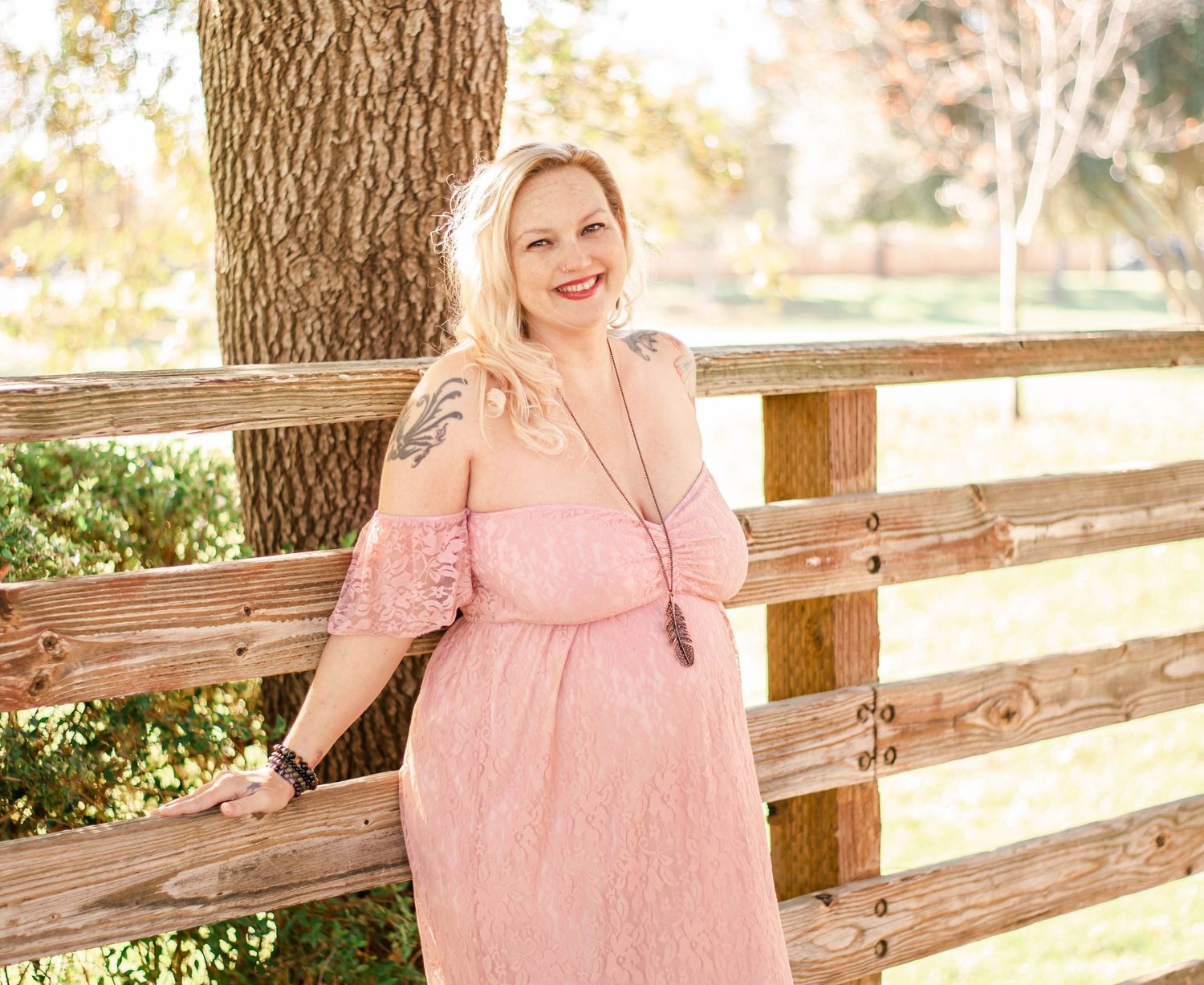 Dreamy maternity photo session in pink lace dress against wooden fence with golden sunlit backdrop.