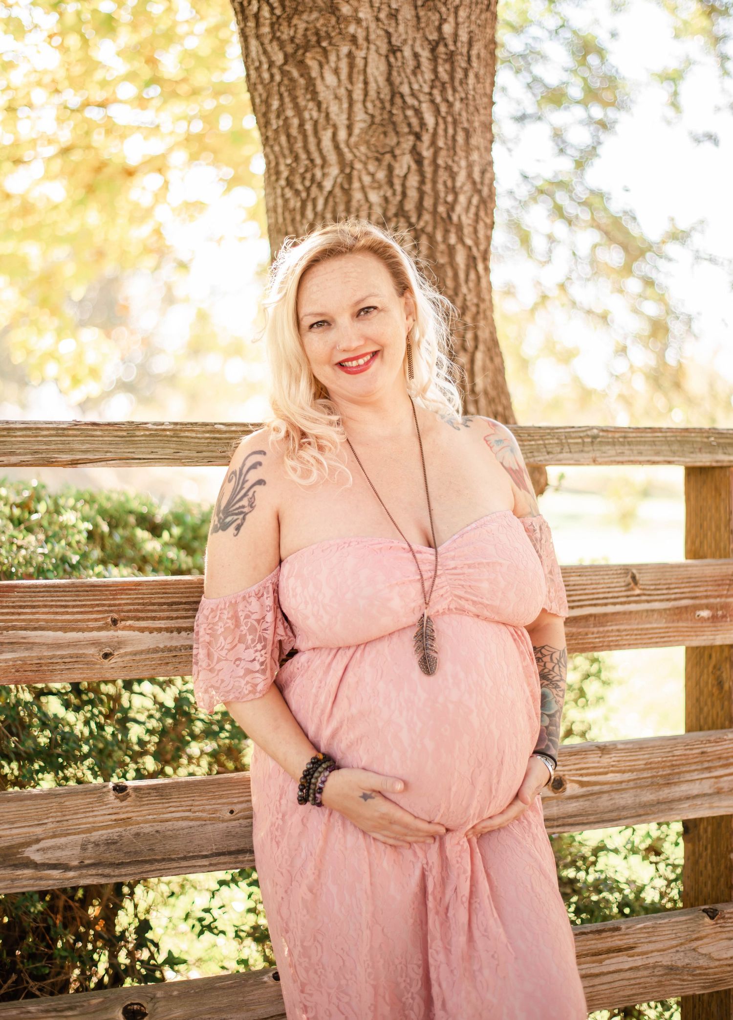 A maternity portrait features a person in pink lace dress posing against a wooden fence.