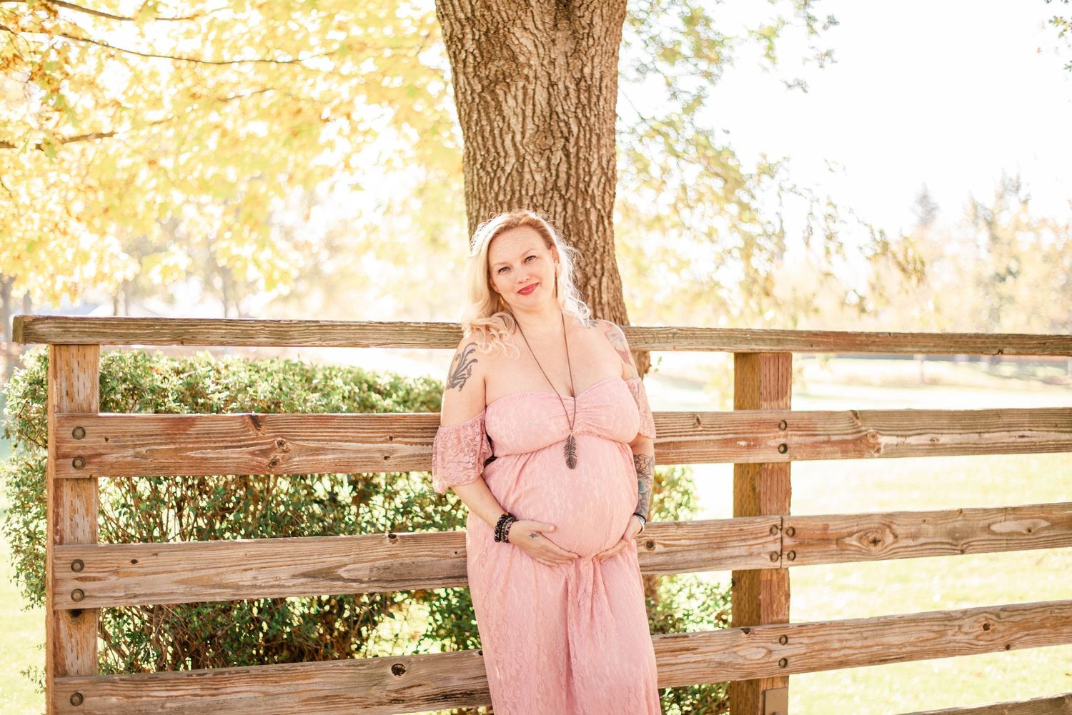 Sunlit maternity photoshoot along wooden fence with pink dress and natural golden lighting.