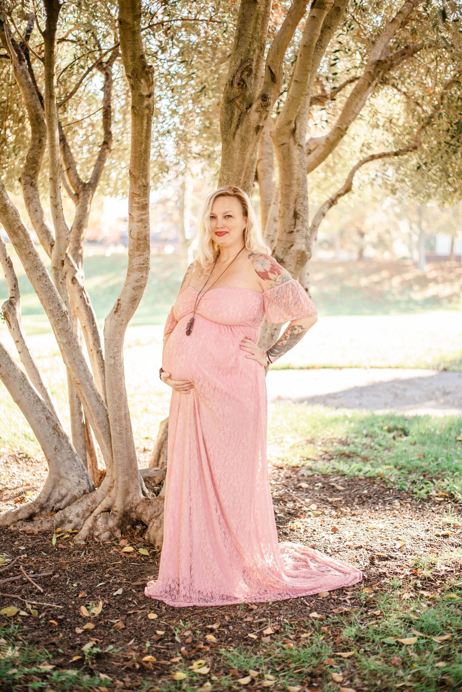 A maternity photoshoot in a wooded area with soft lighting and a flowing pink lace dress.