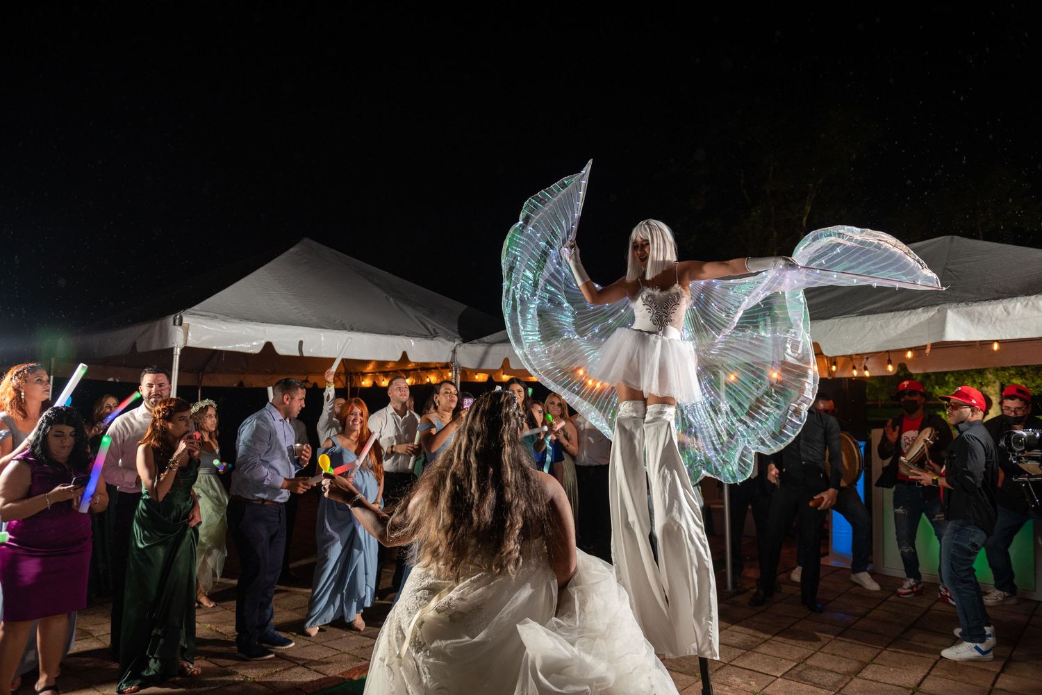 An illuminated angel-like performer entertains wedding guests with glowing wings at an outdoor reception.
