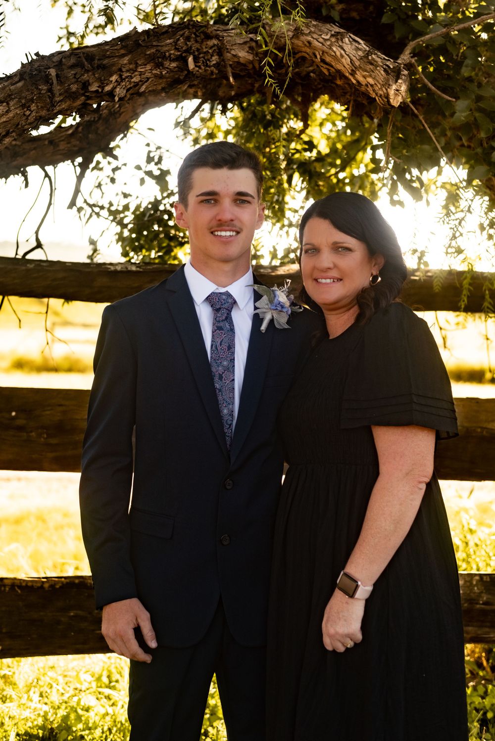 A couple stands together under a rustic wooden arch during sunset in formal attire.