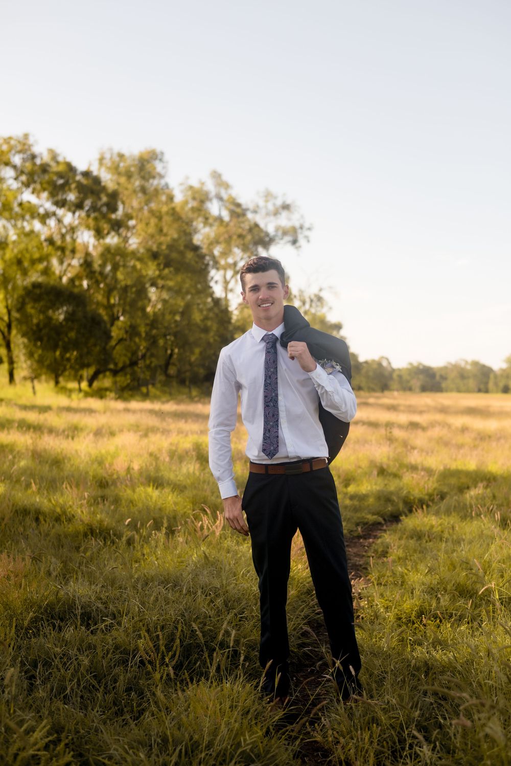 Person in formal attire stands on grassy path at golden hour with trees in background.