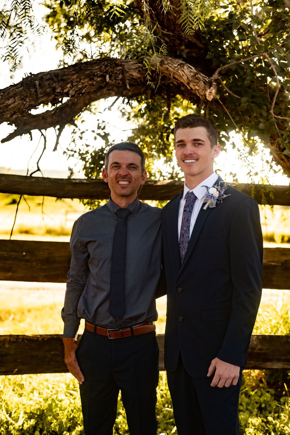 Two people in formal wear stand together under a wooden arch at sunset.