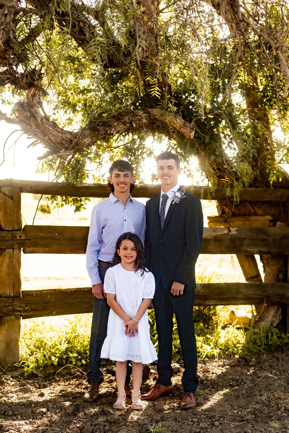 Three people posed together by rustic wooden fence under flowering tree.
