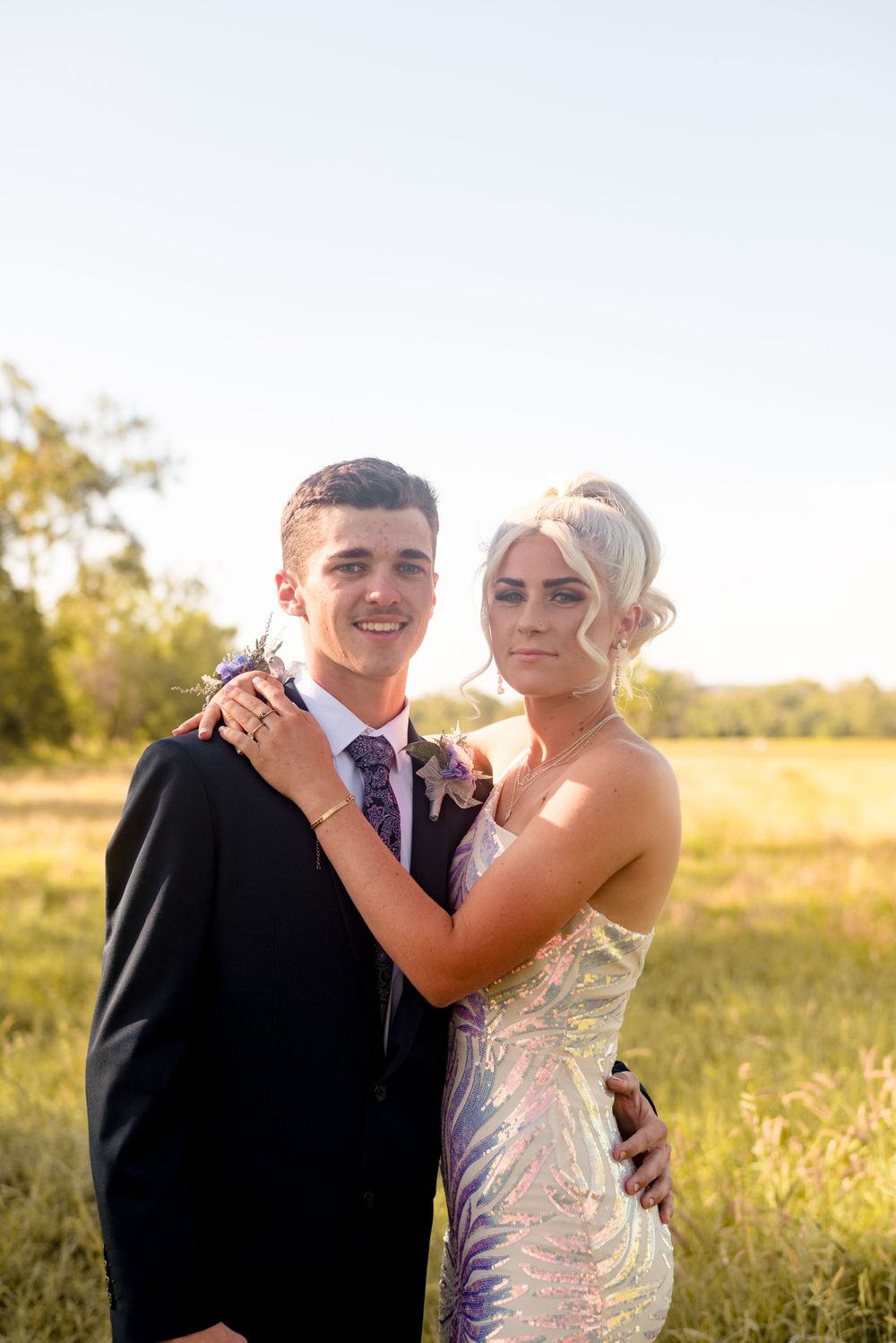 Couple in formal attire embrace during golden hour outdoor photo session in a scenic field.