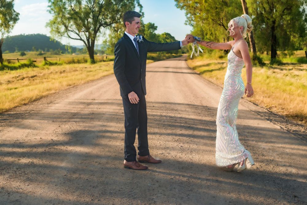 Wedding couple stands facing each other on a rural dirt road surrounded by trees.