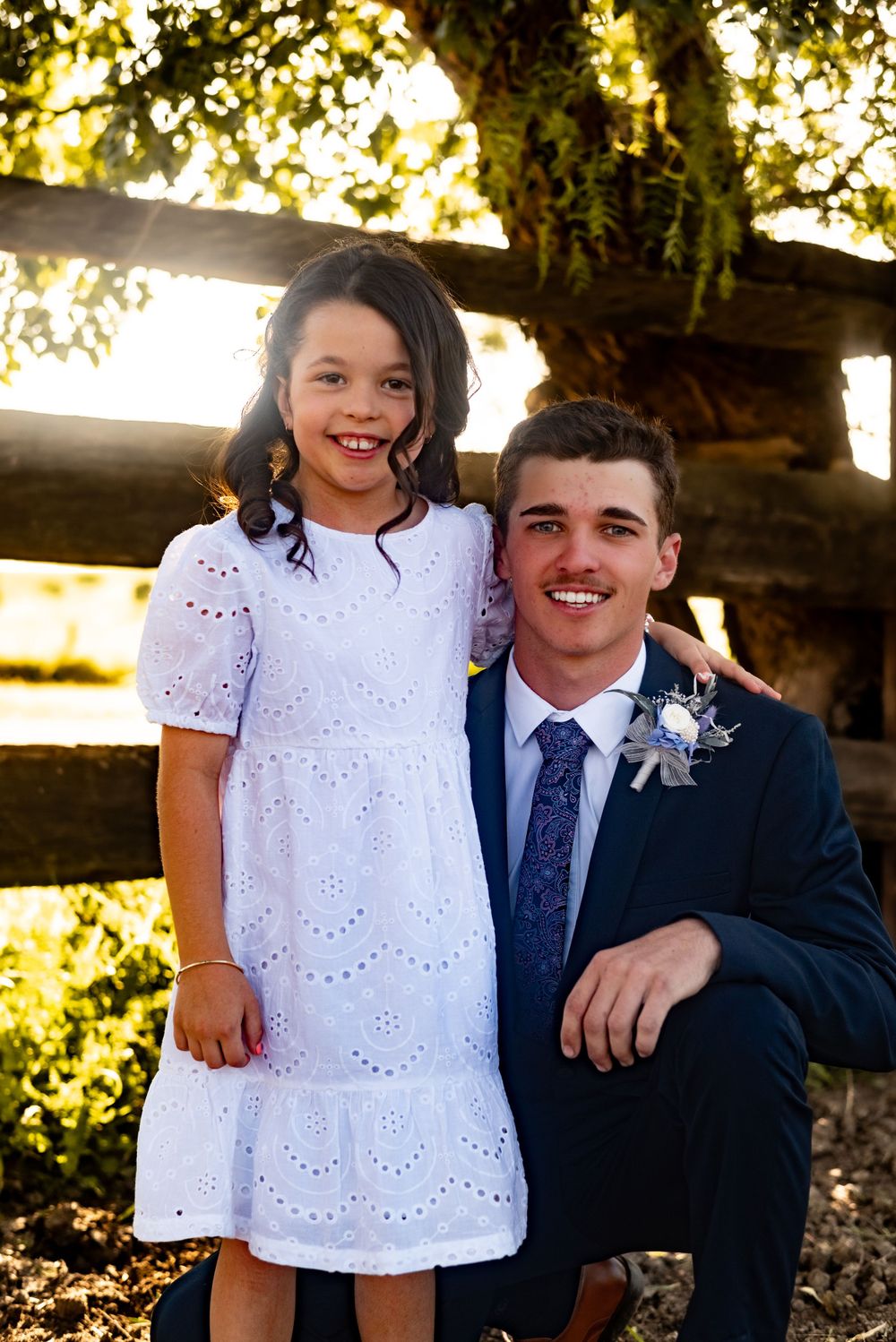 Young girl in white dress poses with formally dressed person by wooden fence at sunset.