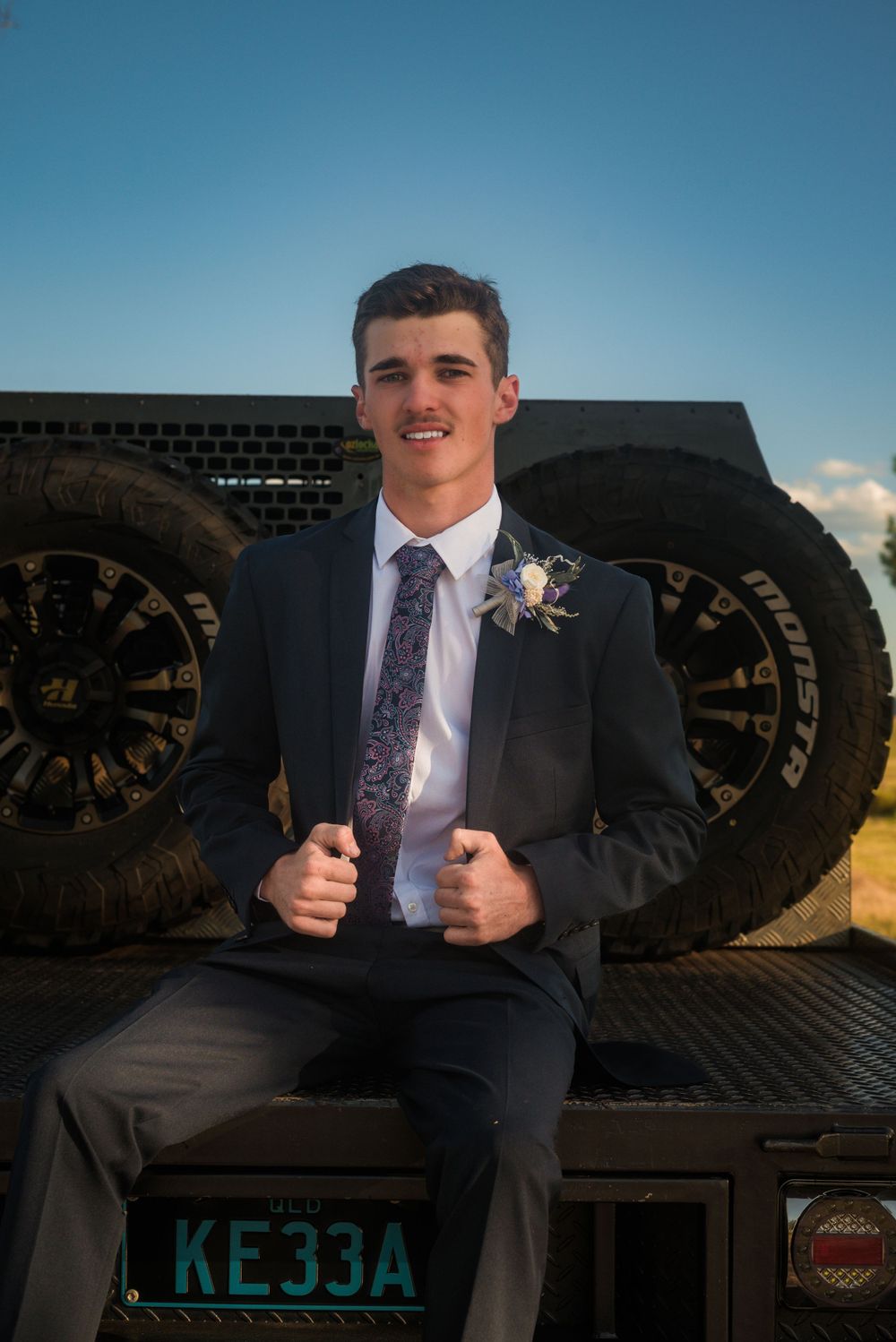 Person in navy suit sits on tailgate of truck with large tires visible.