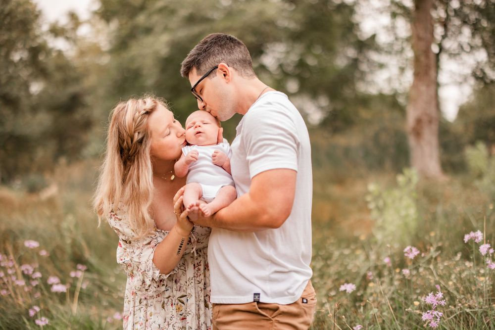 Parents share tender moment with infant in natural outdoor setting with purple wildflowers.