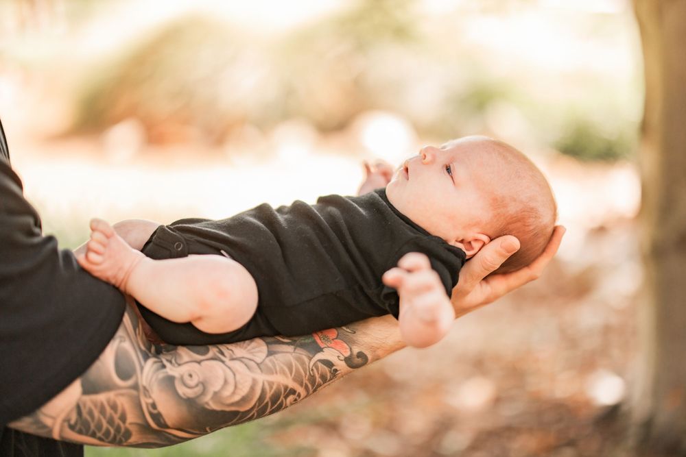 Peaceful sleeping baby in black onesie cradled by tattooed arms outdoors.