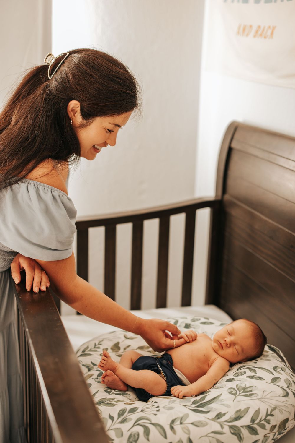 Mother in off-shoulder dress tends to newborn baby lying in dark wooden crib.