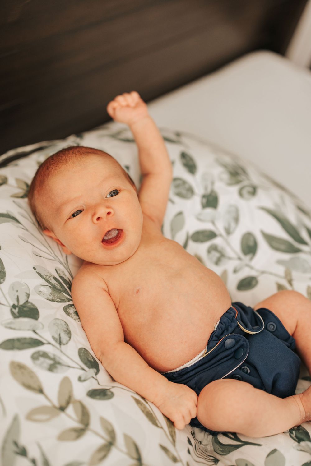 Happy baby in blue diaper stretches and smiles while lying on floral bedding.