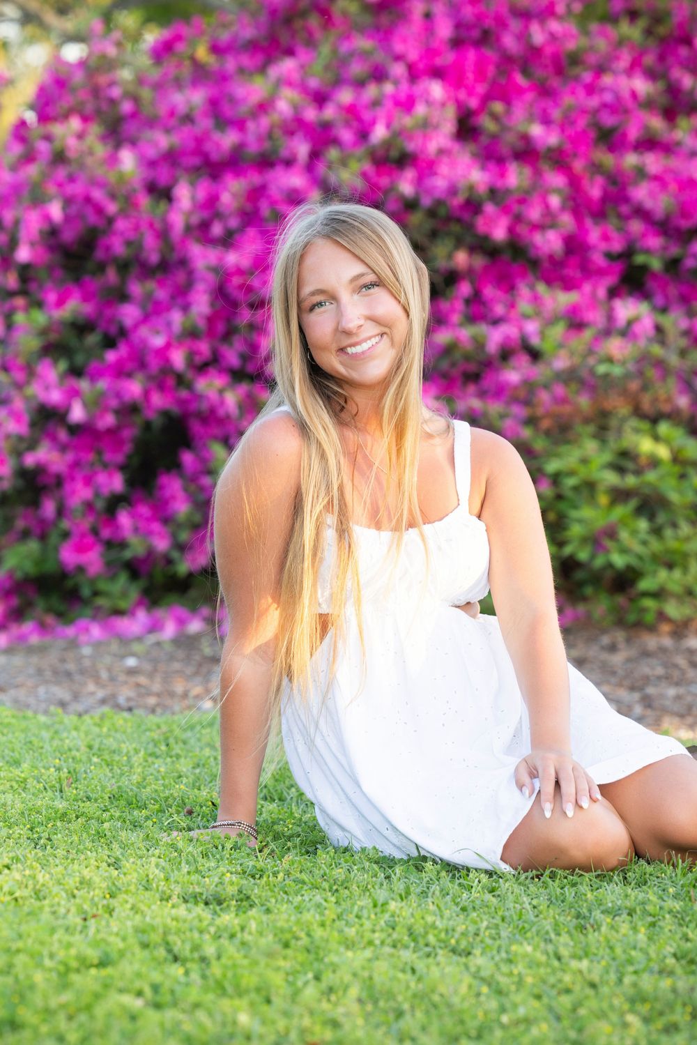 A person in a white sundress sits on grass with vibrant pink bougainvillea flowers blooming in the background.