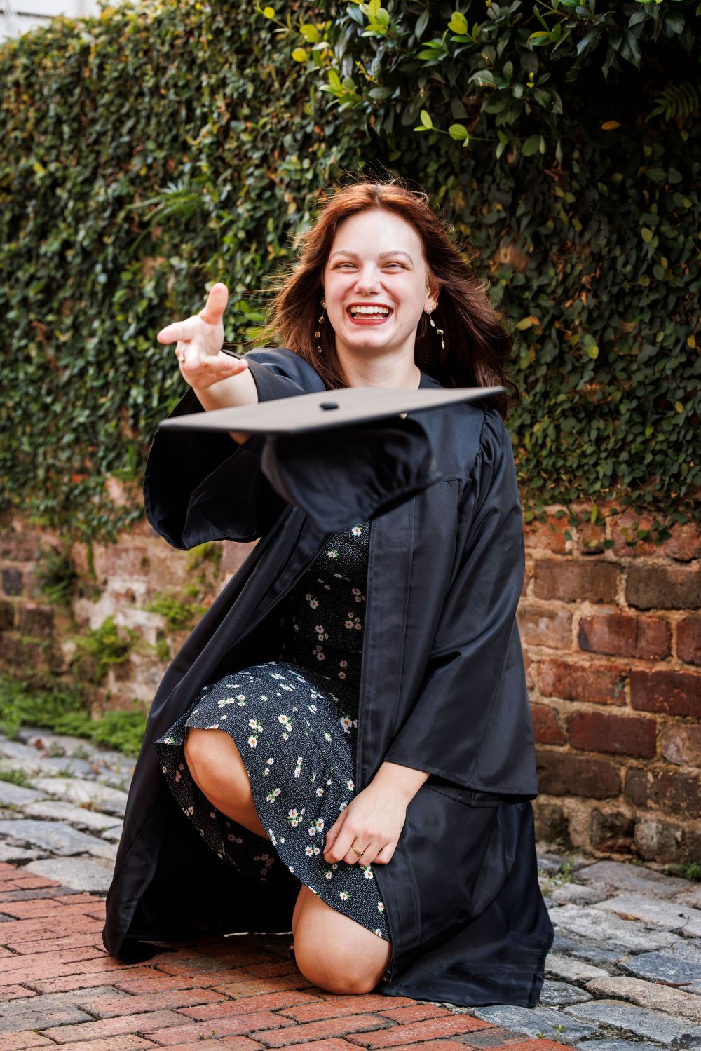 A person in a black coat sits on brick pavement laughing while leaning against a garden wall with foliage.
