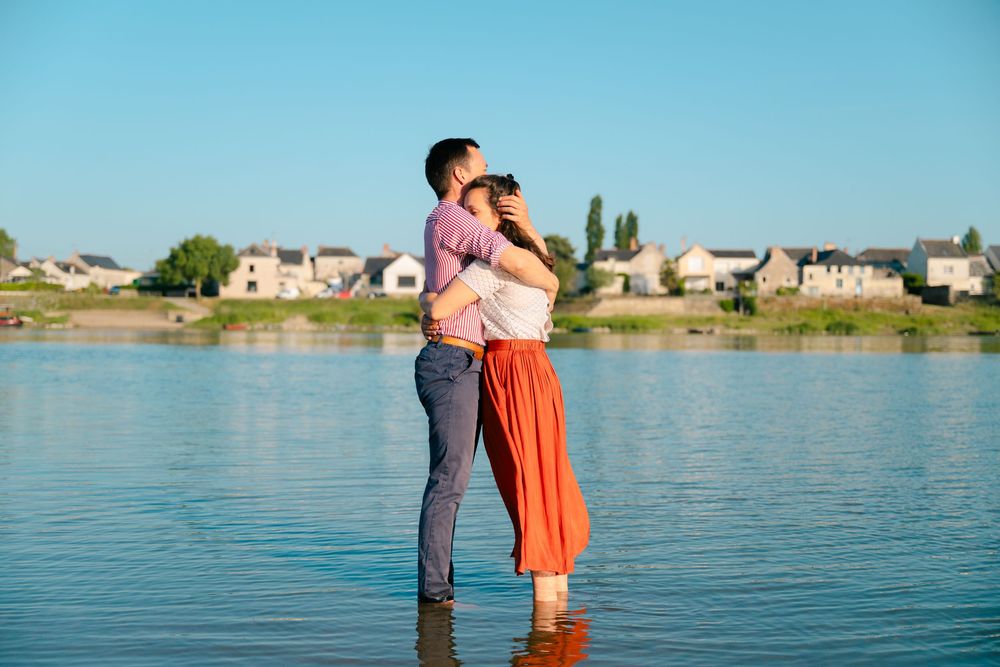 Couple embracing by a tranquil lake on a sunny afternoon with waterfront homes in the background.