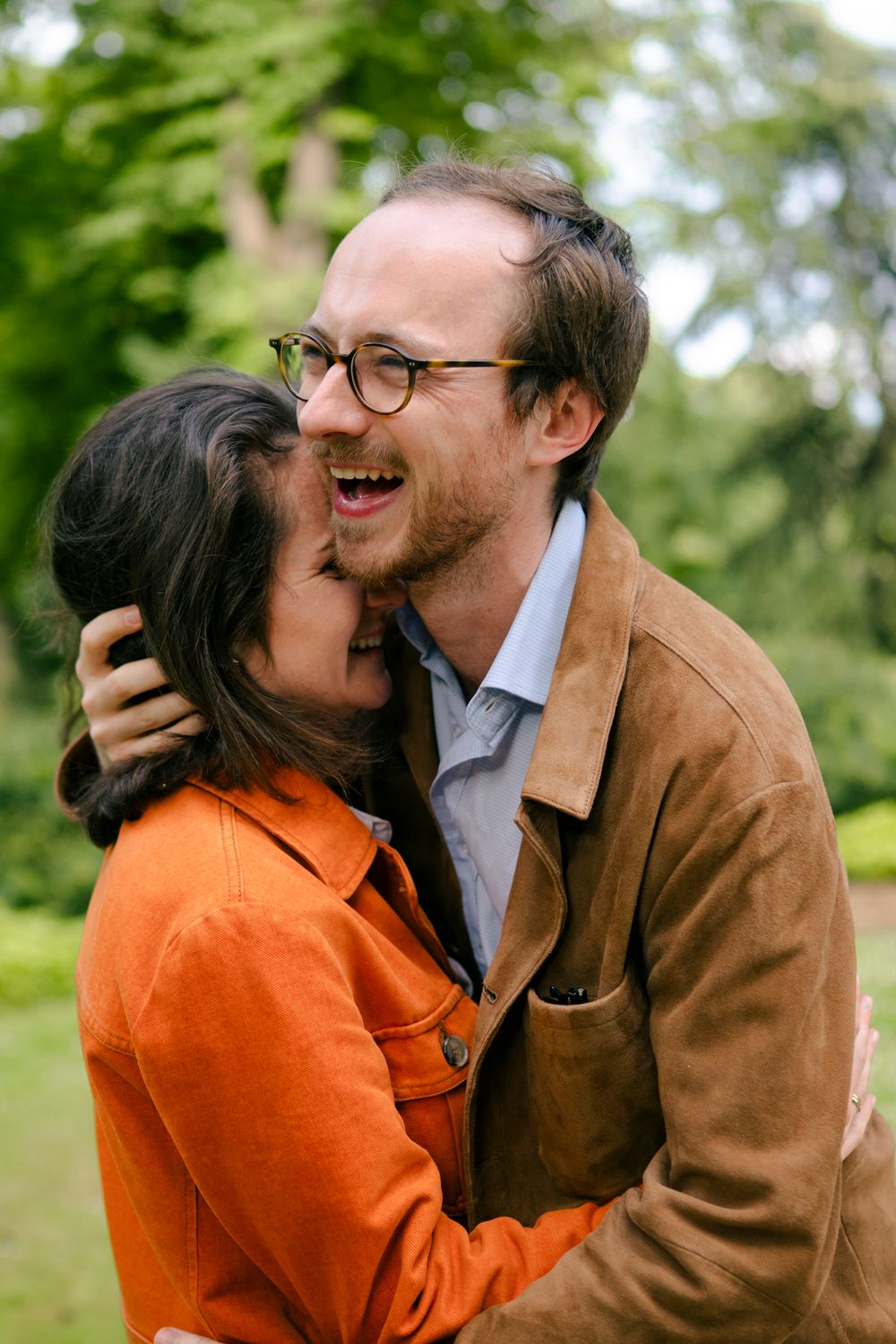A couple shares a joyful embrace outdoors, wearing orange and brown jackets against a green natural background.