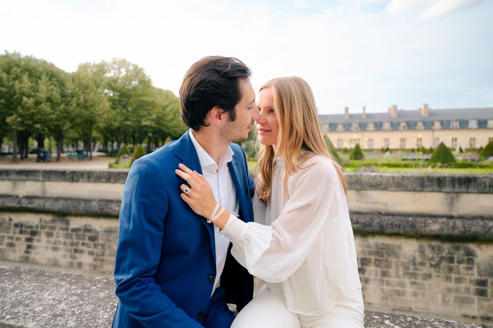 A romantic couple shares an intimate moment by a stone wall in Paris with trees in the background.