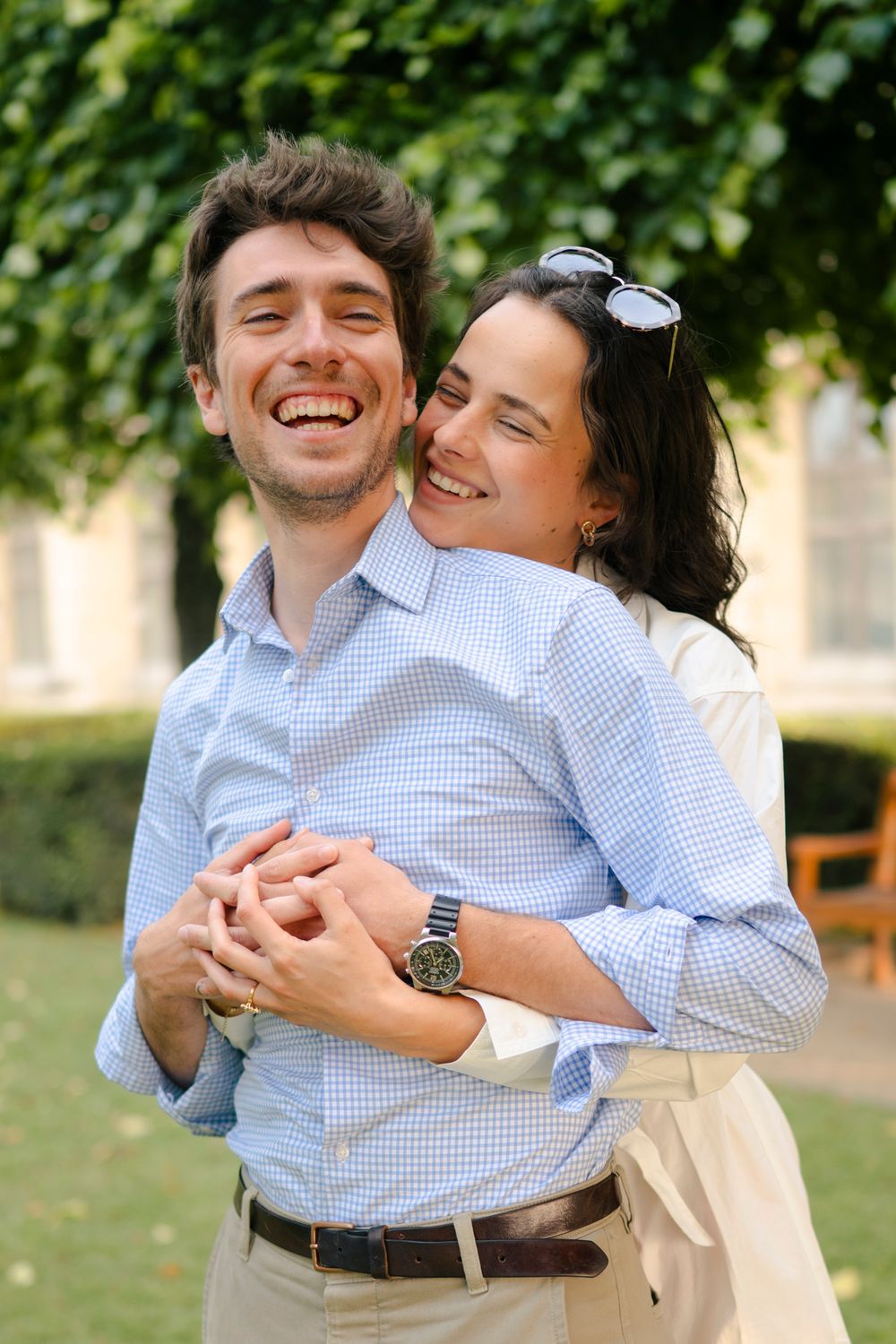 A couple in matching blue gingham shirts embrace and laugh joyfully in a sunny outdoor setting.