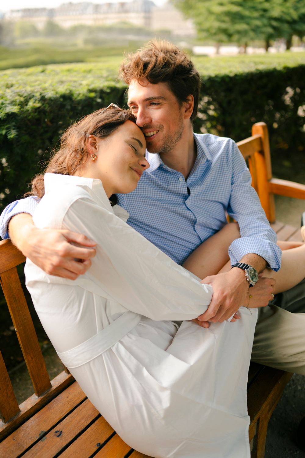A couple shares an intimate moment while sitting together on a wooden bench in a sunny outdoor setting.