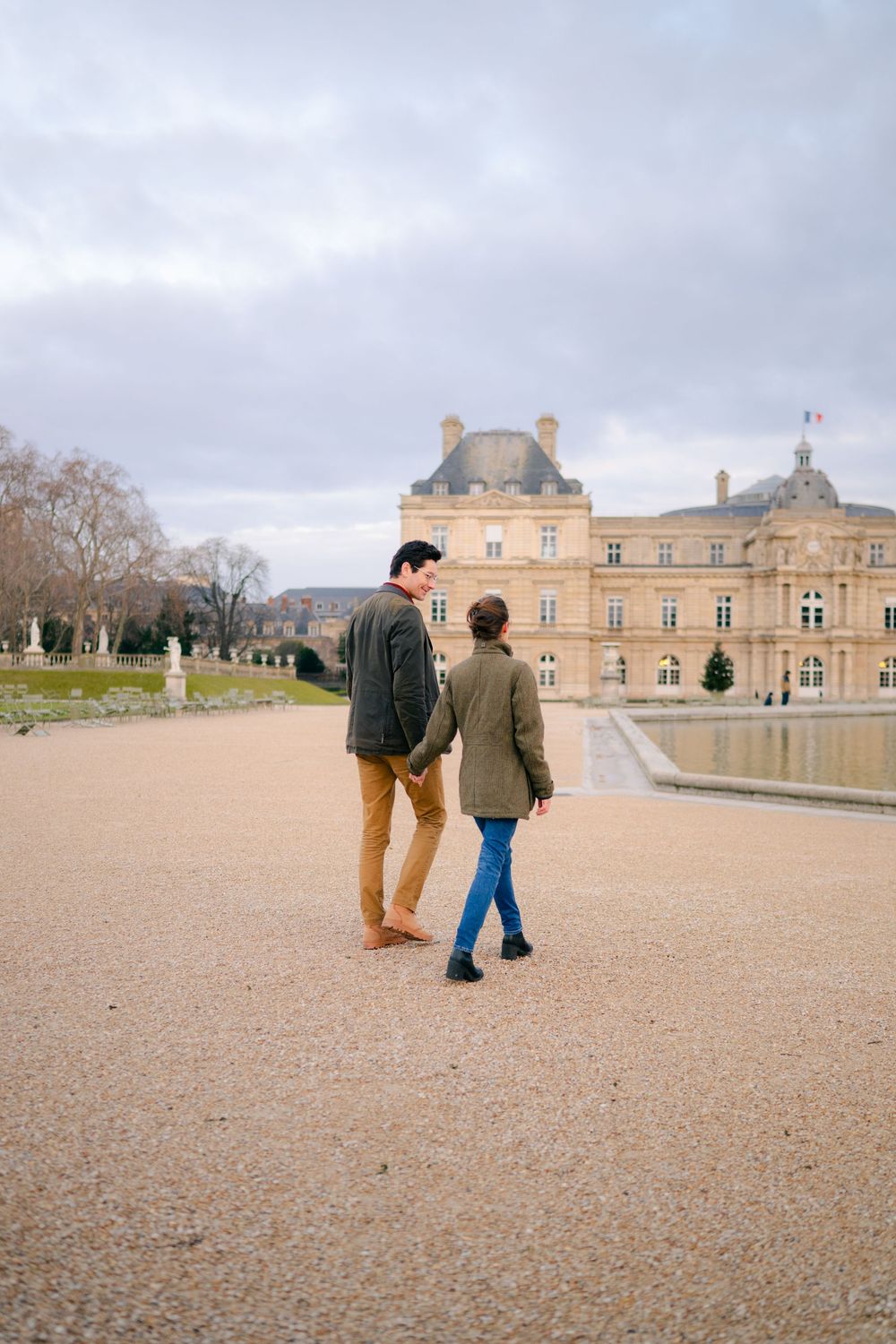 A couple walks along the gravel path in front of the historic Luxembourg Palace in Paris on an overcast day.