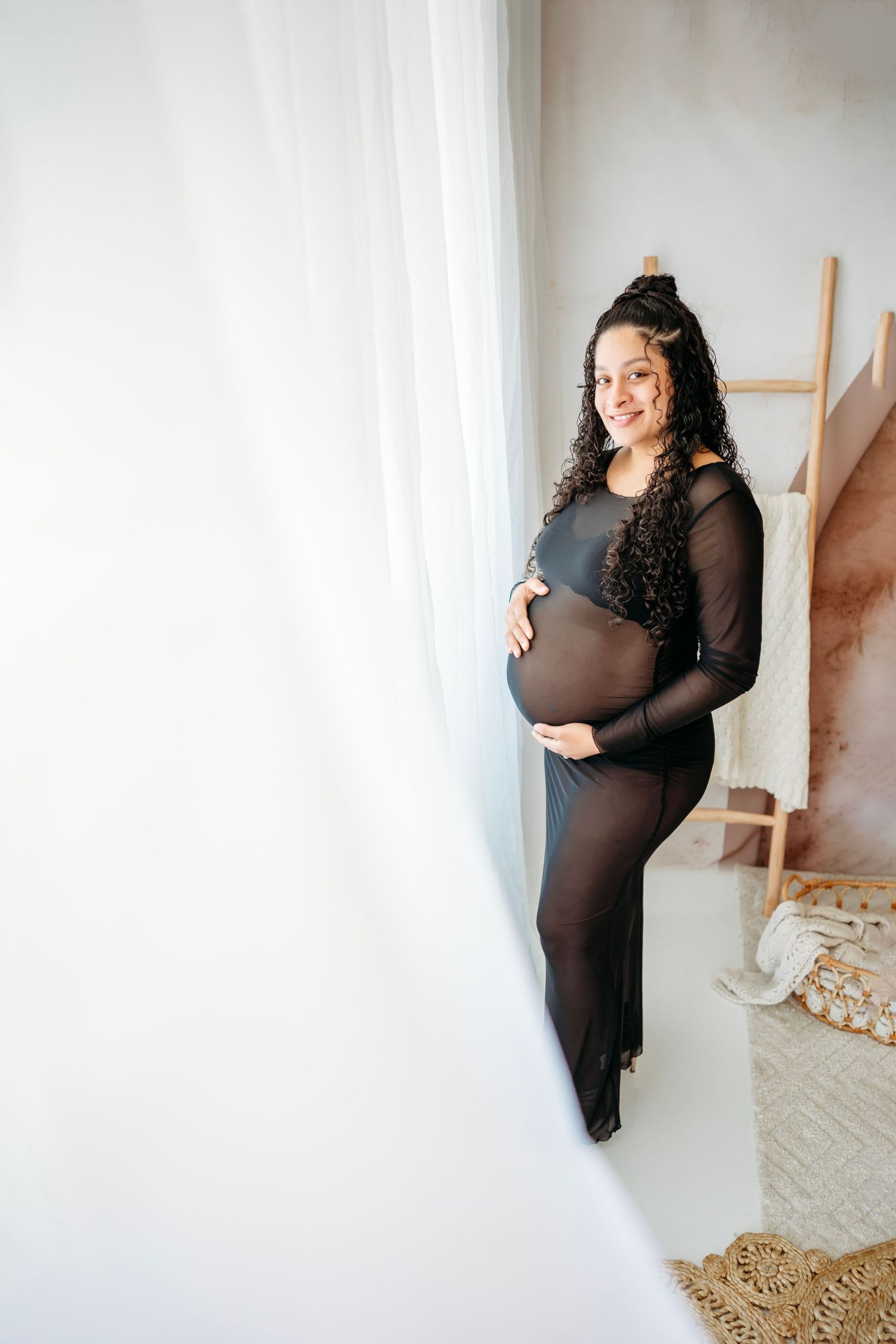 Mom to be wearing a long black dress poses against a white wall in a minimalist interior setting. Maternity Photographer