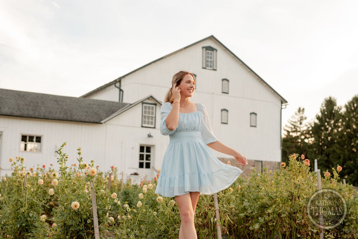 A powder blue dress flows in the breeze as someone poses by a white barn and garden.
