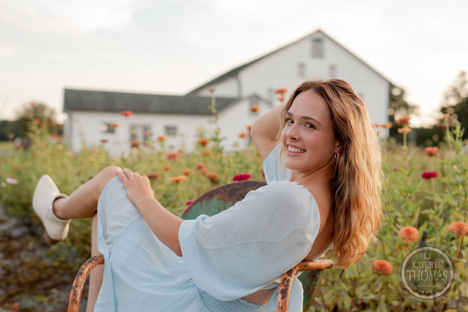 Joyful pose in a light dress while sitting in a wooden chair surrounded by orange flowers.