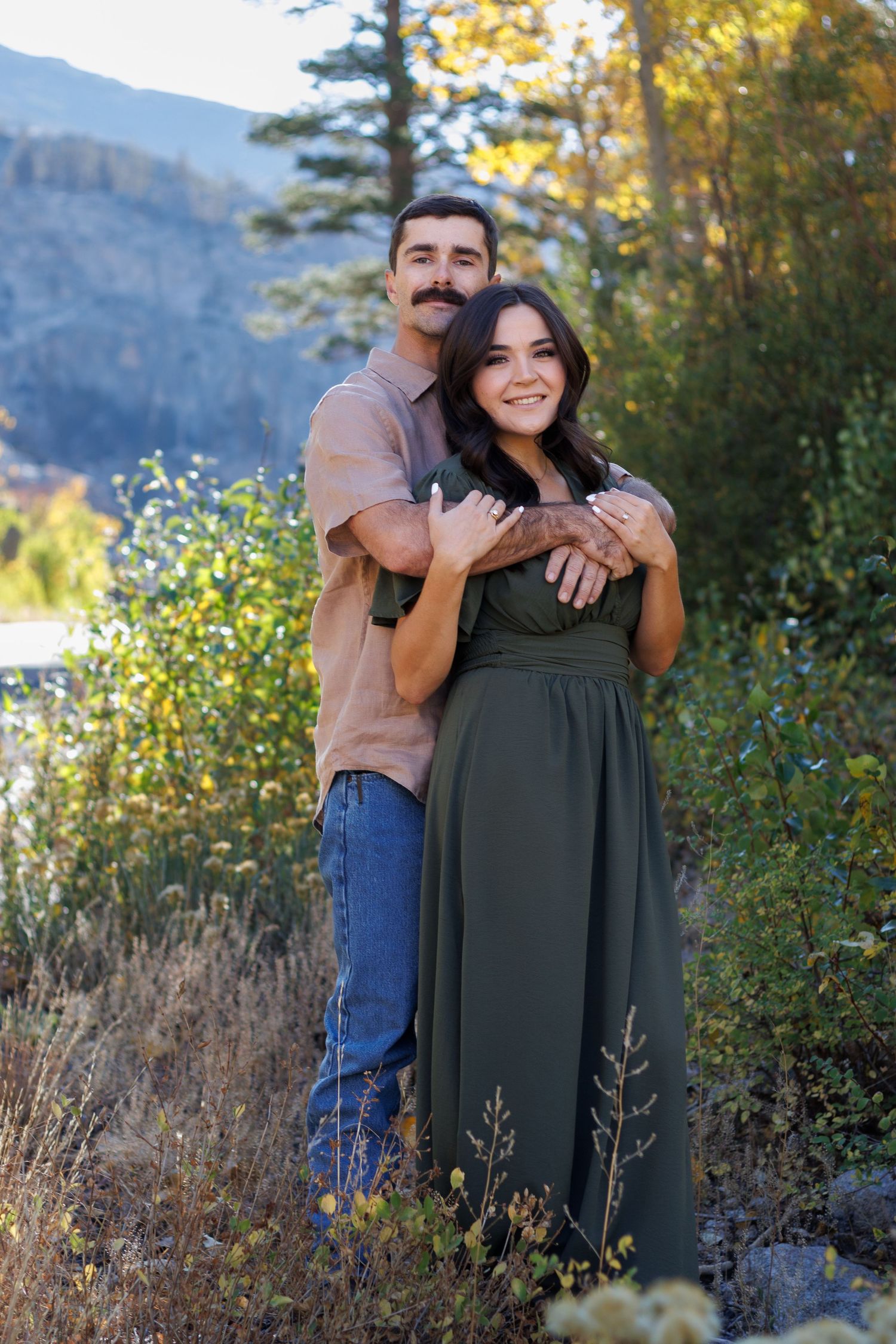 A couple embraces outdoors in front of a mountain landscape on a sunny autumn day.