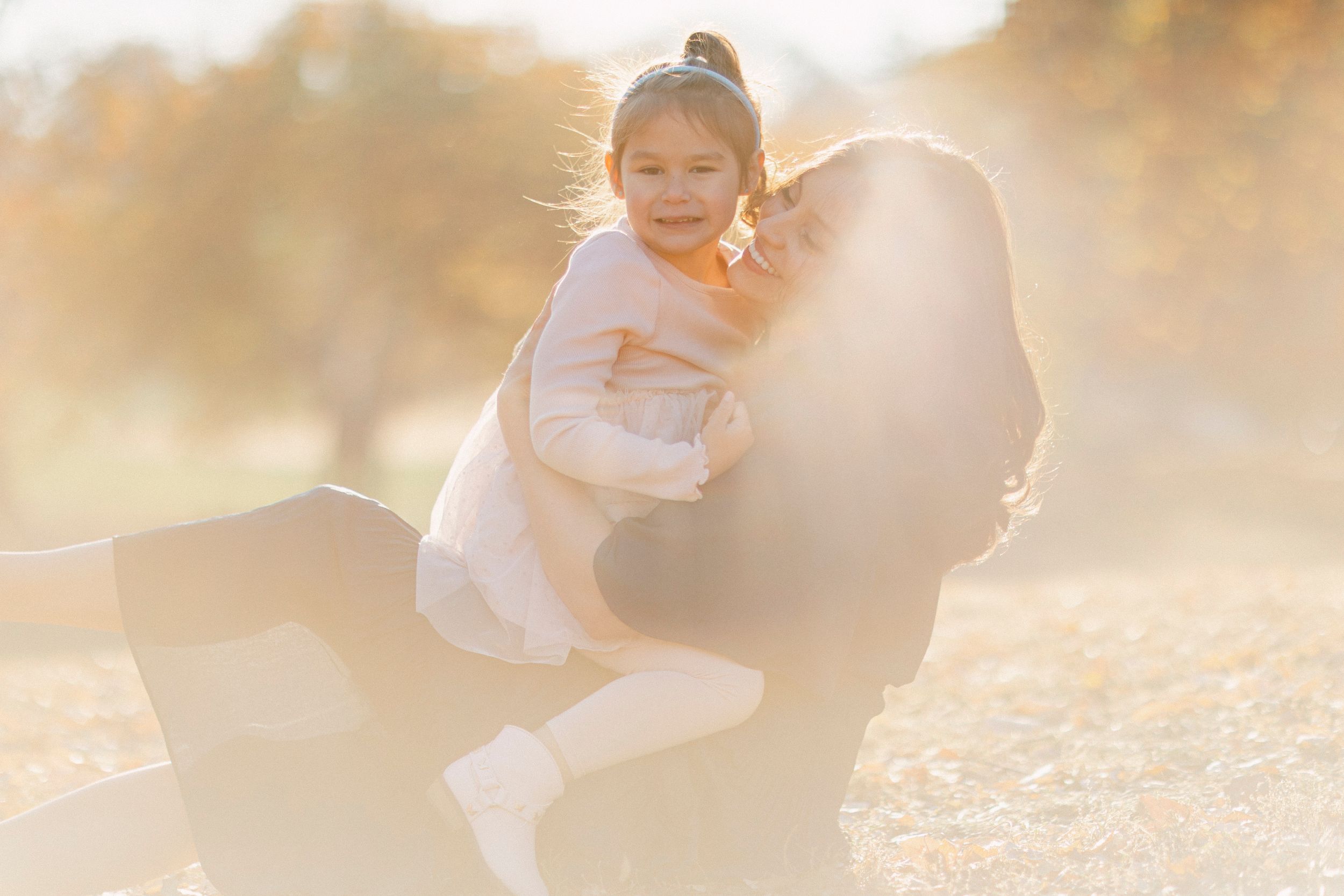 Mother and child share a tender moment in golden sunlit field wearing light colored clothing.