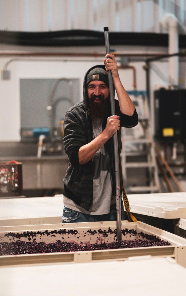 Person in beanie stirring large vat of purple grapes at winery.