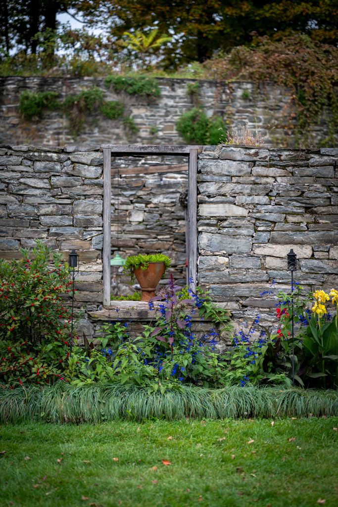 Stone garden wall with wooden frame entrance surrounded by blooming purple flowers and greenery.