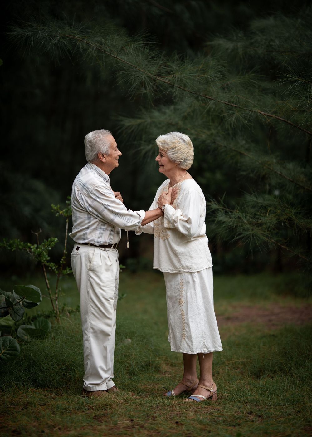 An elderly couple dressed in white share an intimate moment while dancing together in a garden setting.