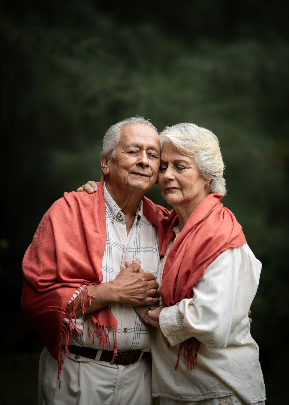 A couple in coral and white embraces in a tender moment against a forest backdrop.